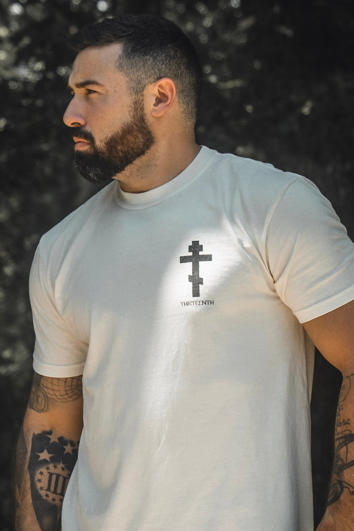 A bearded man stands outdoors wearing the Paid in Full Premium Tee by 13th—a light-colored shirt featuring a black cross and the word THIRTEENTH printed beneath it, symbolizing faith and redemption.
