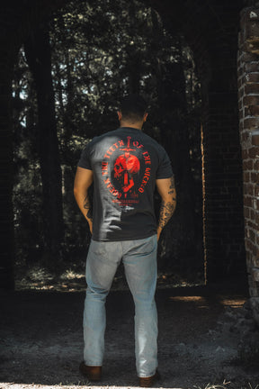 Standing outdoors with their back turned, someone wears the 13th “Break the Teeth Premium Tee”—a black T-shirt featuring a bold red circular graphic—paired with light blue jeans. Framed by brick archways and leafy trees, their thoughtful stance quietly channels Psalm 3:7′s call to “deliver me,” evoking a scene that is both peaceful and resolute.