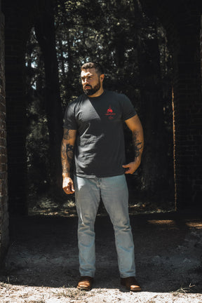 Wearing the 13th “Break the Teeth” Premium Tee and light jeans, a tattooed man stands among brick columns in a shaded outdoor space, trees behind him—a quiet strength in his demeanor as if echoing the defiance found in Psalm 3:7: “Break the teeth of the wicked.”.