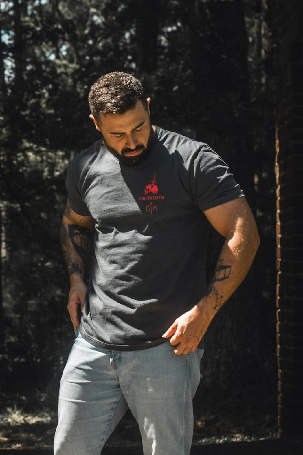 Wearing the Break the Teeth Premium Tee by 13th, a tattooed man with a beard and short hair stands outdoors in sunlight, pairing the grey T-shirt with light blue jeans. As he gazes downward with hands near his pockets, his thoughtful expression seems to echo Psalm 3:7’s plea, “deliver me.”.