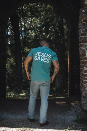 A man faces away from the camera, standing outdoors near a brick archway with trees in the background. He is wearing the 13th brand “Jesus Saves Premium Tee - Seafoam,” which references John 3:16 and its message of everlasting life.