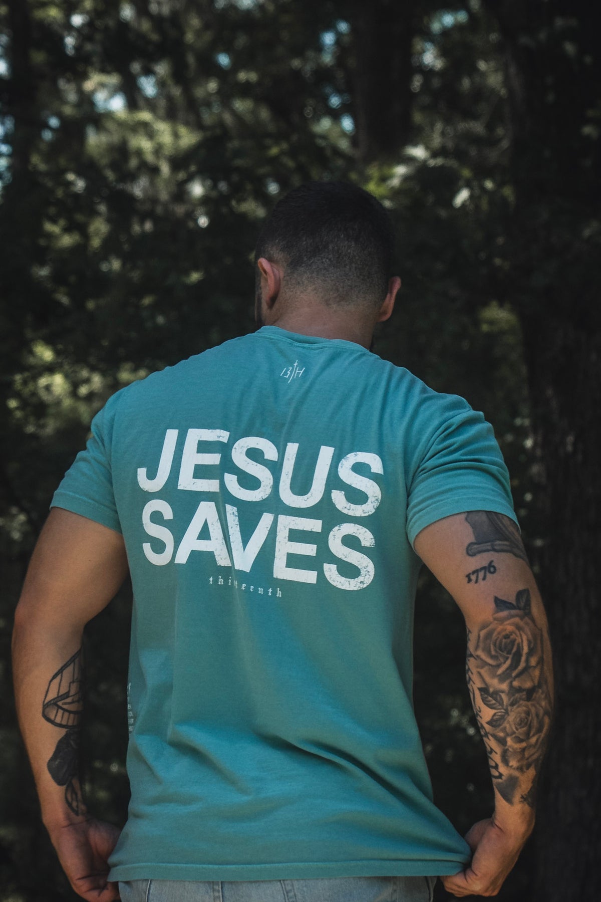 A man with tattoos stands outdoors wearing the 13th Jesus Saves Premium Tee in Seafoam, featuring “JESUS SAVES” printed in bold white letters on the back—a reference to the promise of everlasting life in John 3:16.