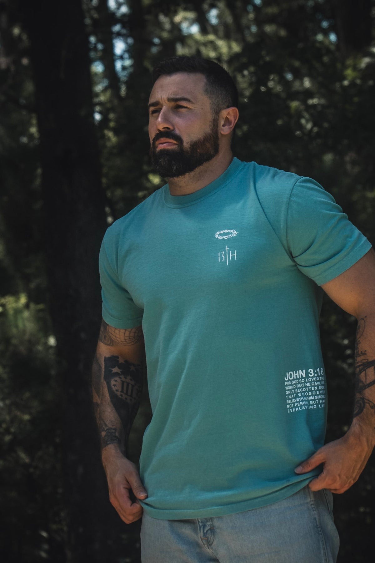 A bearded man stands outdoors amidst trees and greenery, wearing the 13th Jesus Saves Premium Tee in Seafoam. The T-shirt features white text and graphics on the chest and lower side, highlighting the Bible verse John 3:16 about everlasting life. He pairs it with light jeans, completing a relaxed, peaceful look.