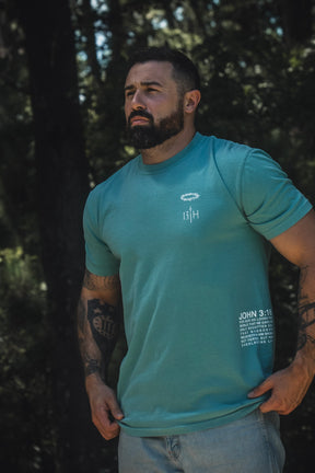 A bearded man stands outdoors amidst trees and greenery, wearing the 13th Jesus Saves Premium Tee in Seafoam. The T-shirt features white text and graphics on the chest and lower side, highlighting the Bible verse John 3:16 about everlasting life. He pairs it with light jeans, completing a relaxed, peaceful look.
