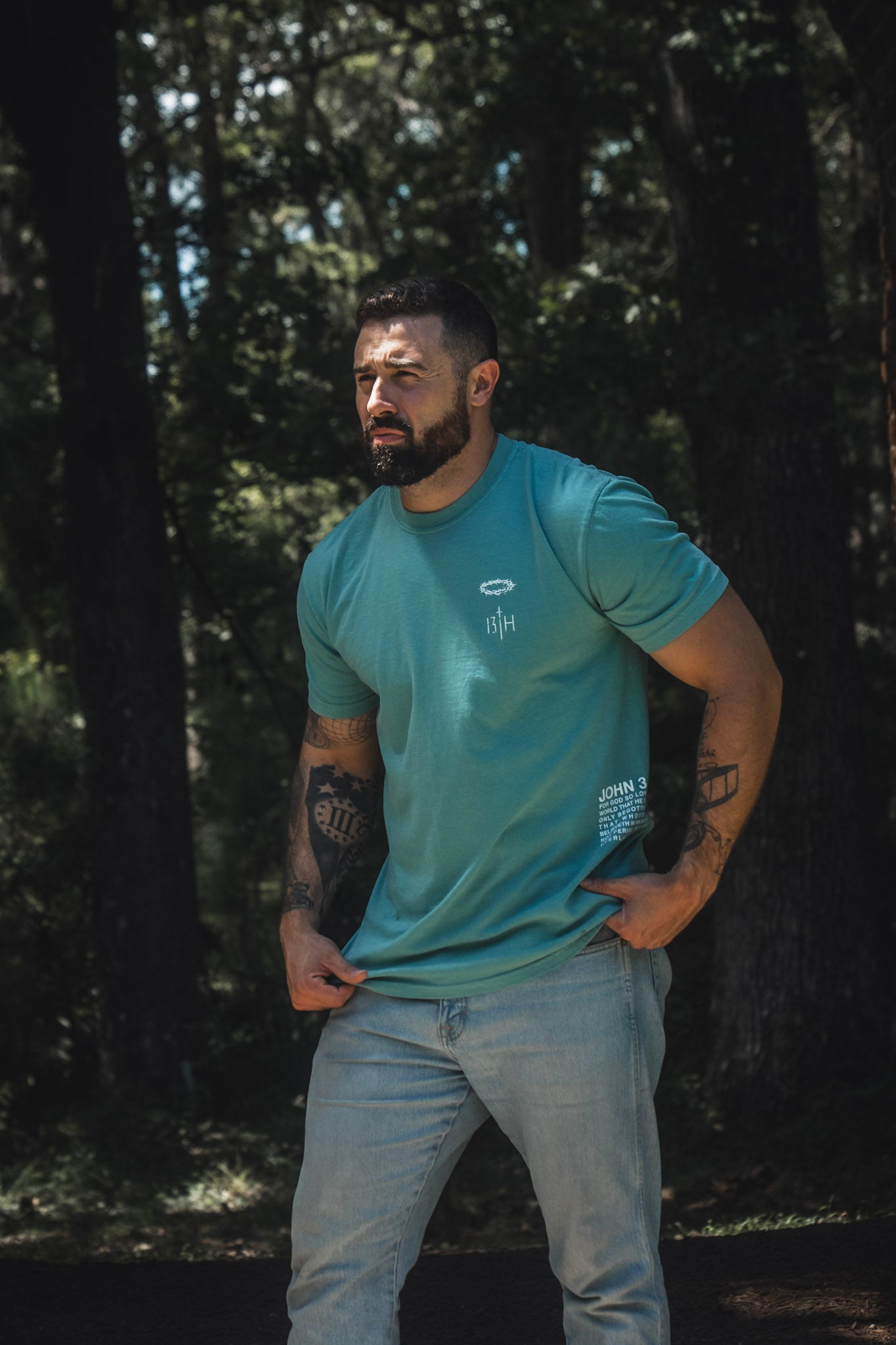 A bearded man stands outdoors in a wooded area, wearing the 13th Jesus Saves Premium Tee in Seafoam with jeans, thoughtfully holding the hem of his shirt as he reflects on the message of everlasting life found in John 3:16.