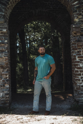 A man with tattoos wears the 13th Jesus Saves Premium Tee in Seafoam and light jeans, standing under a brick archway bathed in sunlight and trees—a scene that captures the hopeful promise of everlasting life found in John 3:16.