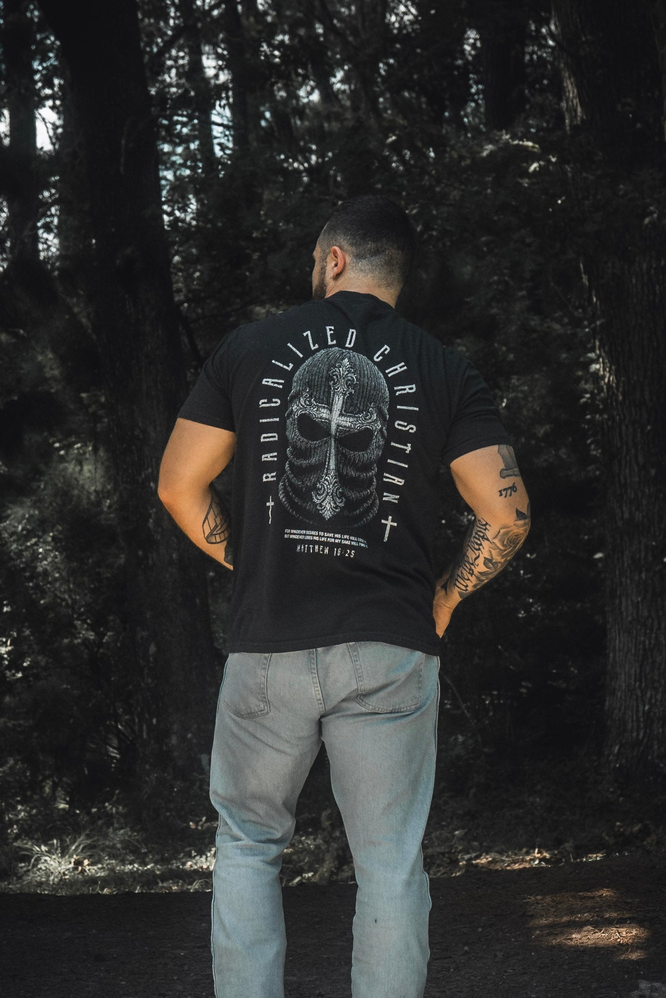 A man stands in a forest, facing away from the camera, wearing light blue jeans and the 13th Radicalized Christian Premium Tee—a black shirt featuring a helmet graphic and the Bible verse Matthew 16:25: “For whoever wants to save his life will lose it, but whoever loses his life for me will find it.”.