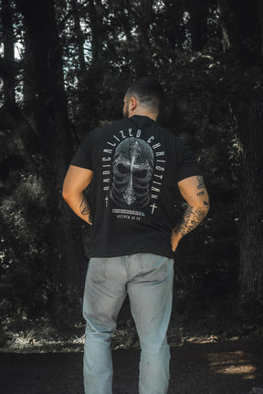 A man stands in a forest, facing away from the camera, wearing light blue jeans and the 13th Radicalized Christian Premium Tee—a black shirt featuring a helmet graphic and the Bible verse Matthew 16:25: “For whoever wants to save his life will lose it, but whoever loses his life for me will find it.”.