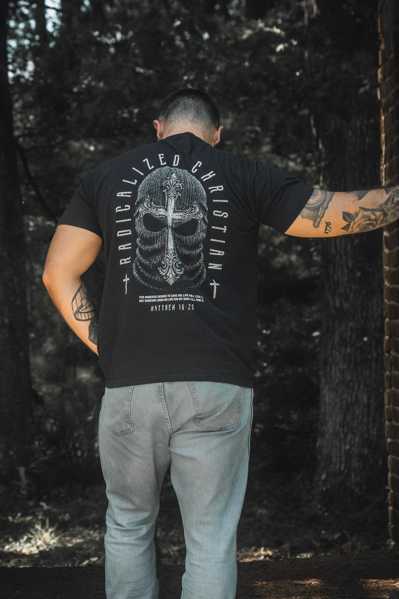 A person stands outdoors with their back to the camera, wearing the 13th Radicalized Christian Premium Tee in black, which displays “Radicalized Christian,” an illustrated helmet graphic, and the Bible verse Matthew 16:25 on the back.