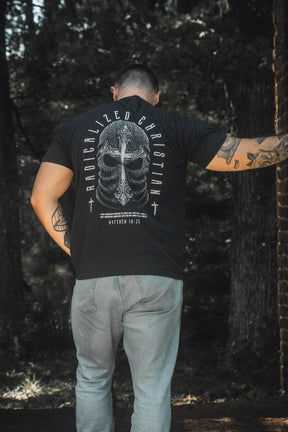 A person stands outdoors with their back to the camera, wearing the 13th Radicalized Christian Premium Tee in black, which displays “Radicalized Christian,” an illustrated helmet graphic, and the Bible verse Matthew 16:25 on the back.