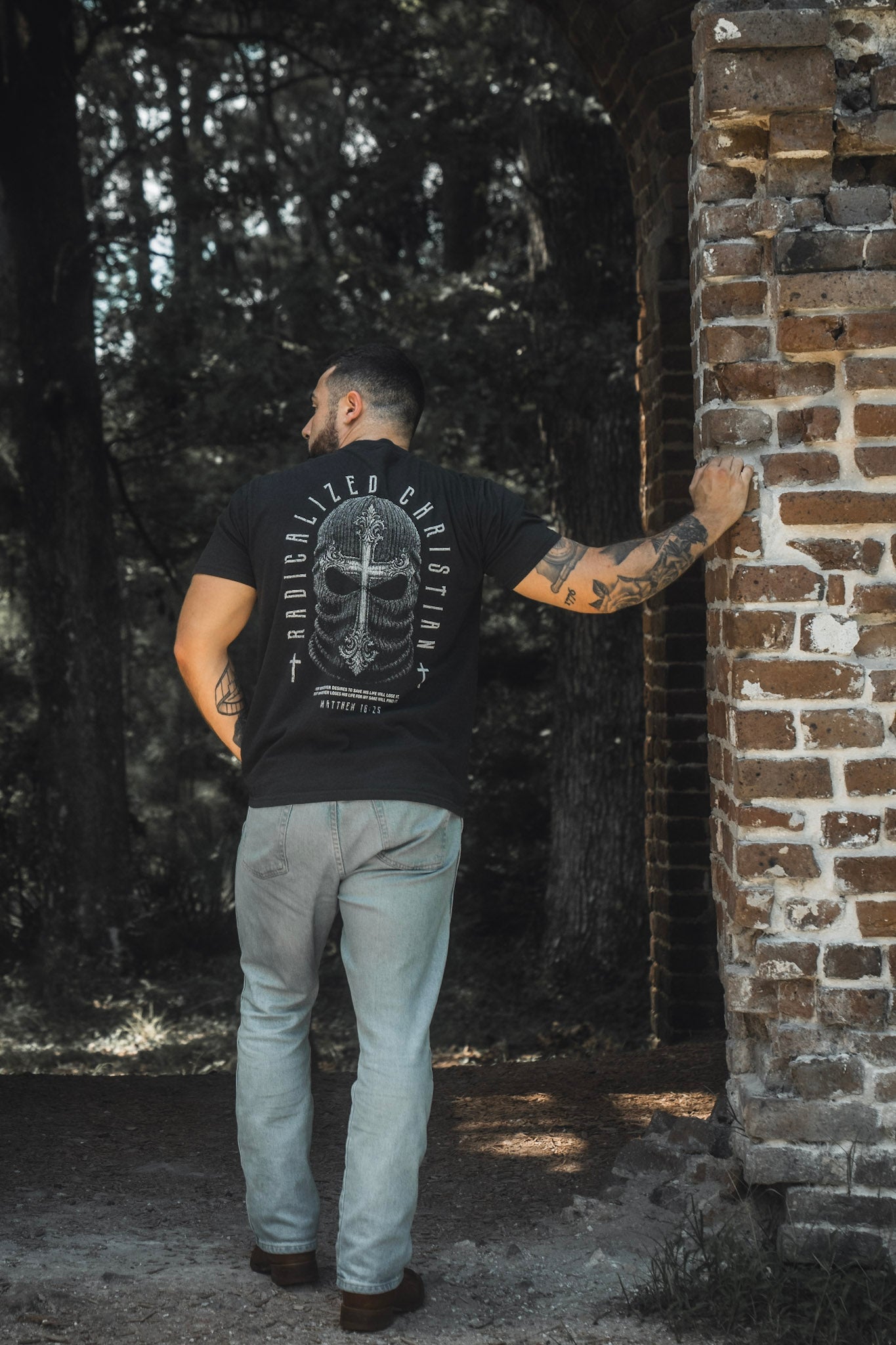 A man with tattoos, dressed in jeans and the 13th Radicalized Christian Premium Tee—a black graphic t-shirt featuring a Bible verse—stands outdoors with one hand resting on a brick wall, facing away from the camera.