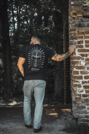 A man with tattoos, dressed in jeans and the 13th Radicalized Christian Premium Tee—a black graphic t-shirt featuring a Bible verse—stands outdoors with one hand resting on a brick wall, facing away from the camera.