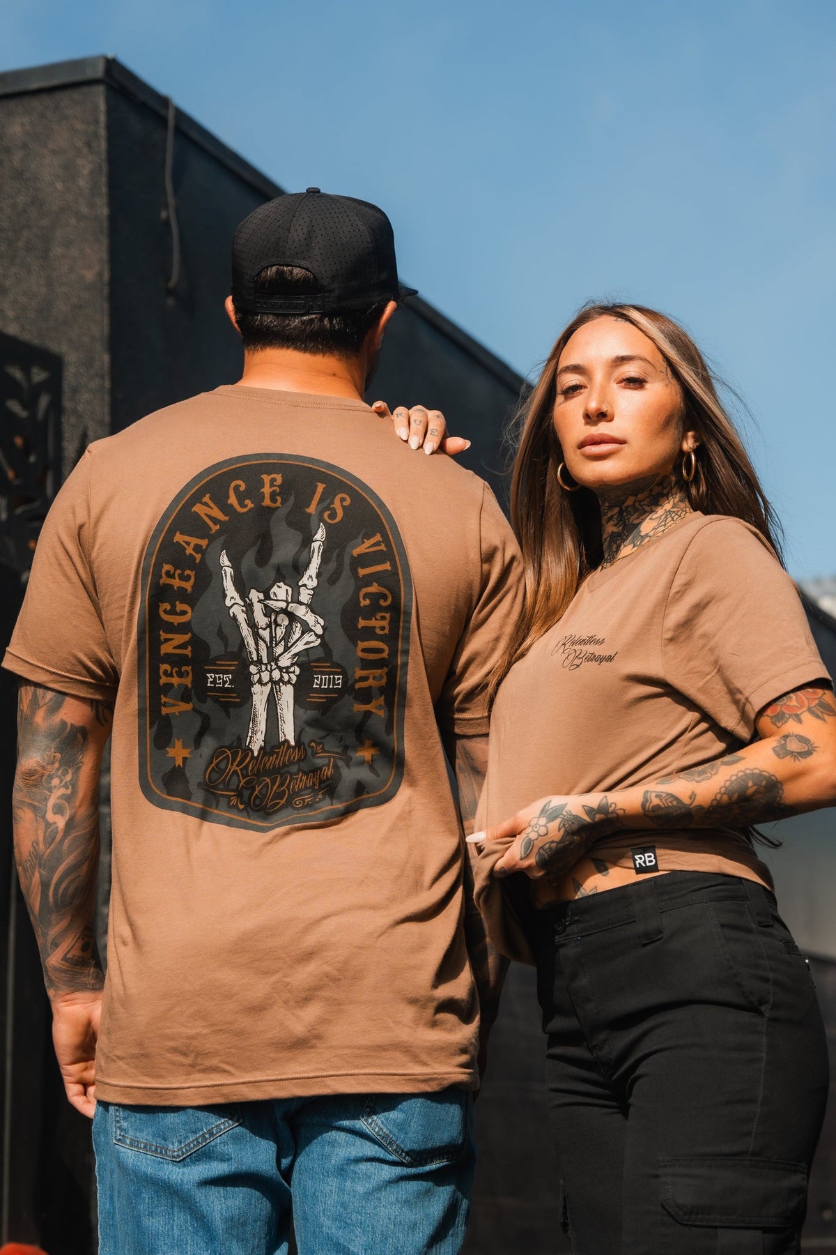 A tattooed man in a cap and a Relentless Betrayal Vengeance Camel T-shirt stands next to a tattooed woman in similar style. Their confident pose under the clear blue sky symbolizes resilience and empowerment.
