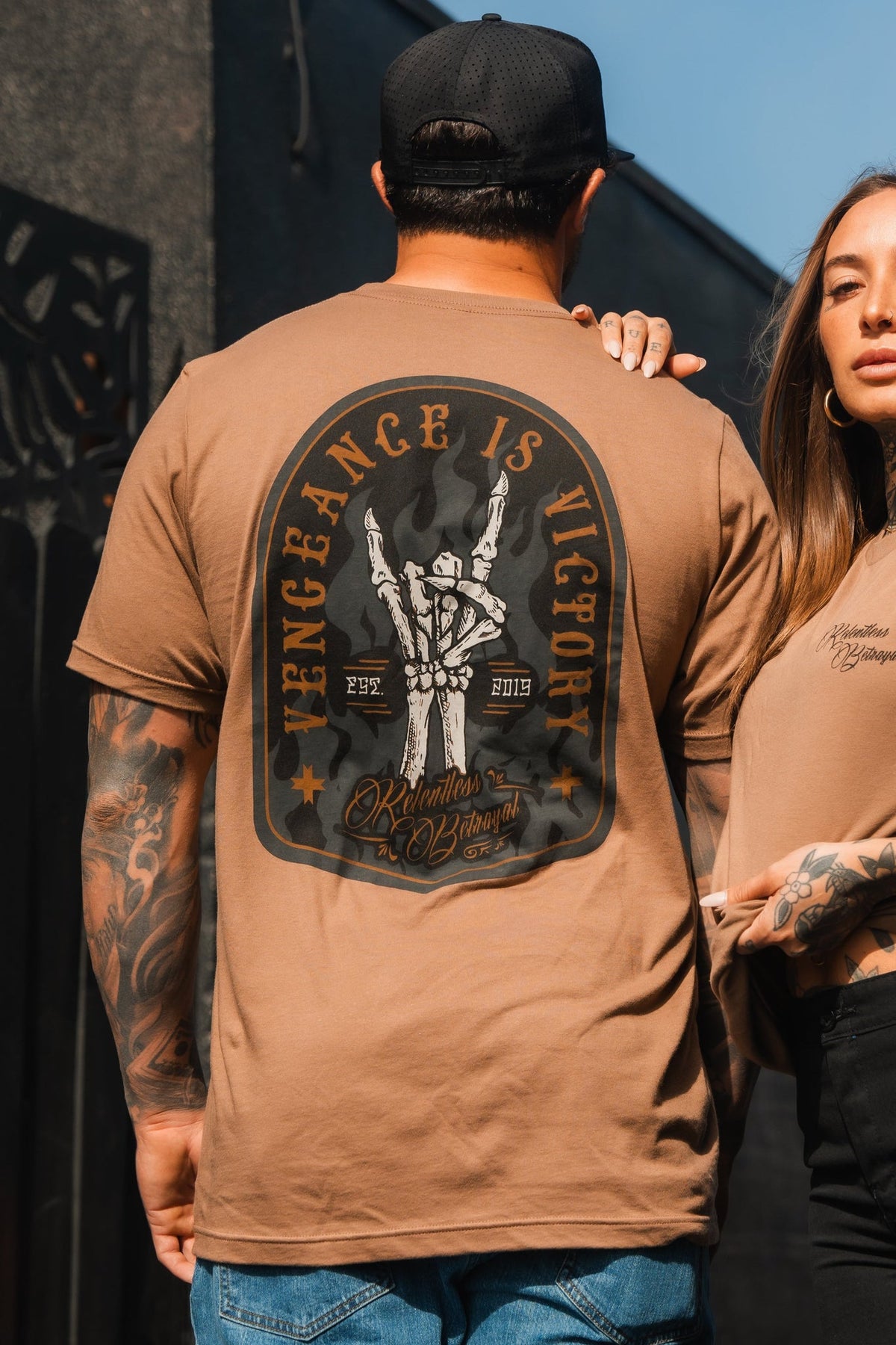 A man in a brown Vengeance Camel T-Shirt from Relentless Betrayal stands with a woman in matching apparel. They exhibit tattoos symbolizing resilience, set against a dark outdoor backdrop, embodying empowerment through their bold clothing choice.