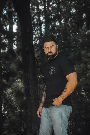 A bearded man stands in a forest, sporting the 13th Radicalized Christian Premium Tee in black and light jeans, one hand tucked into his pocket. Tattoos adorn his arms, including the Bible verse Matthew 16:25—a reminder that whoever wants to save his life will lose it, but whoever loses his life for a greater purpose will find it.