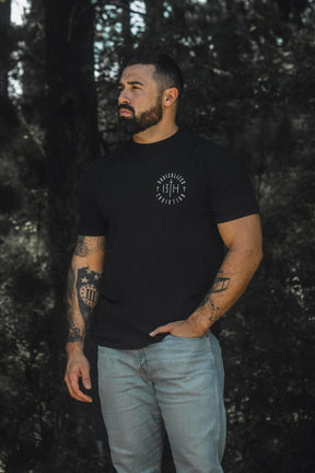 A tattooed, bearded man stands outdoors against a forest backdrop in dim natural light, dressed in the Radicalized Christian Premium Tee by 13th and light jeans. With one hand in his pocket and looking contemplatively to the side, he seems to reflect on the message of Matthew 16:25—a verse about discovering what it truly means to save your life.