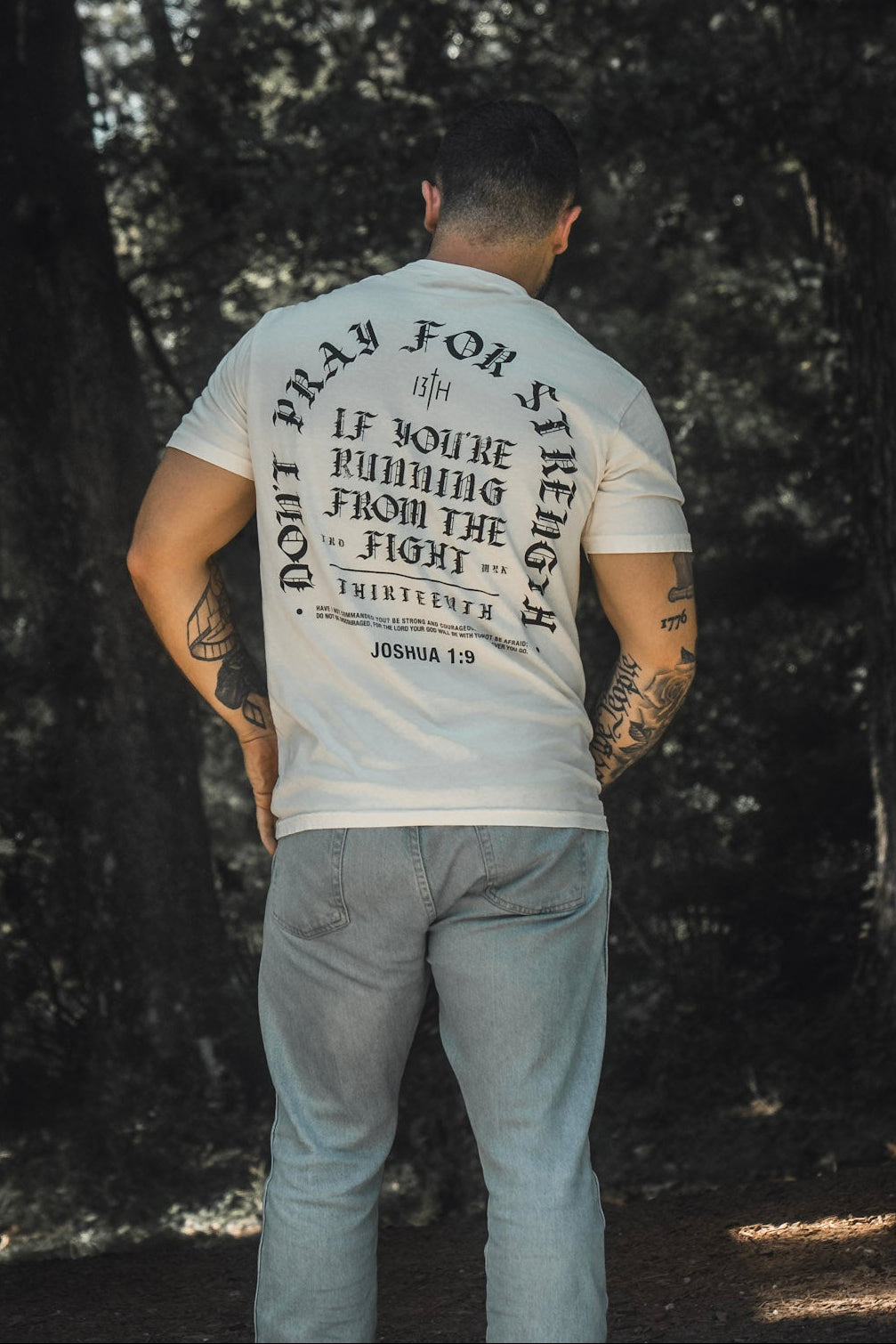 A man stands outdoors with his back to the camera, wearing the Be Strong Premium Tee from 13th, which features a printed quote from Joshua 1:9, surrounded by trees and partial brickwork.