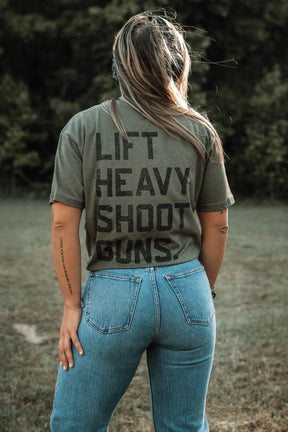 A person with long hair, wearing a green Tango Charlie Apparel "Lift Heavy. Shoot Guns." Next Level CVC shirt and blue jeans, stands outdoors with their back to the camera. Trees and grass are visible in the background.