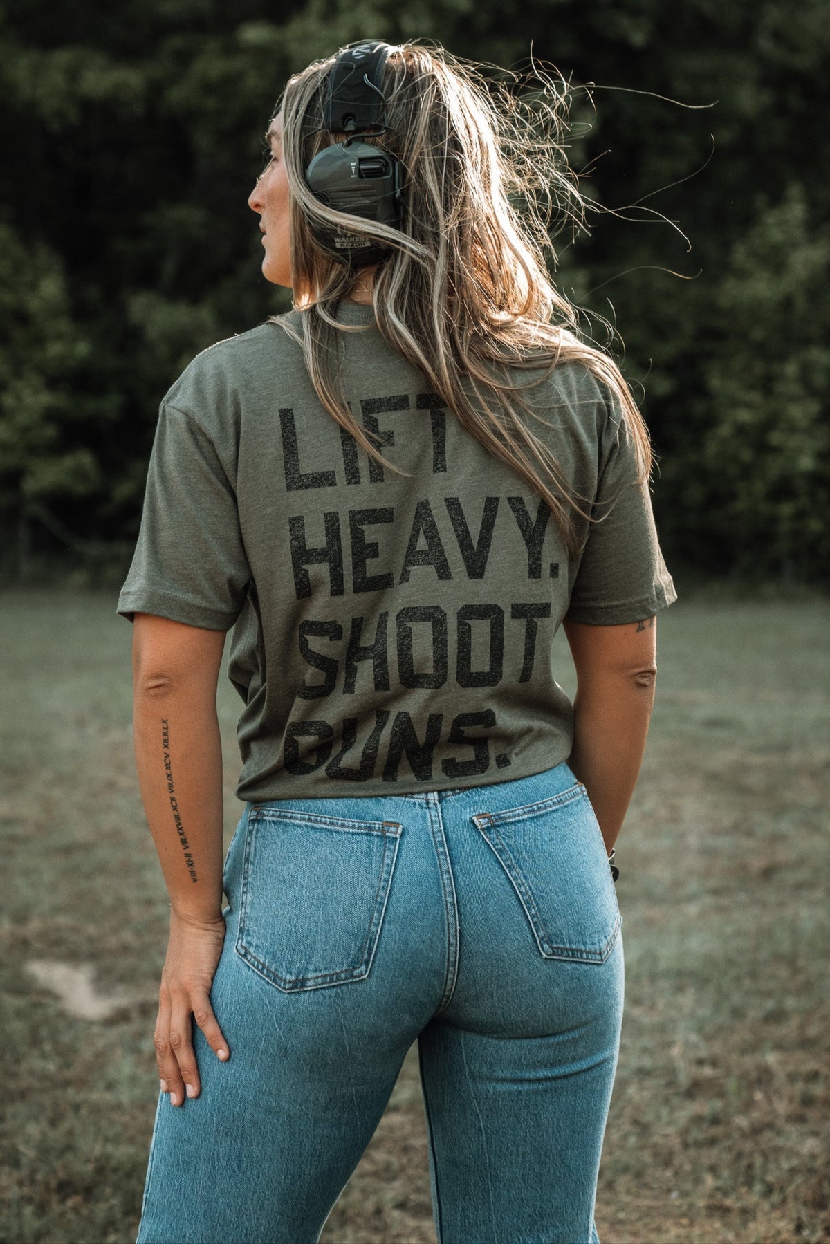 A woman with tattooed arms stands outdoors in jeans, wearing a green "Lift Heavy. Shoot Guns." shirt by Tango Charlie Apparel, made of polyester and combed ringspun cotton, with ear protection on her head, facing away toward trees and grass.