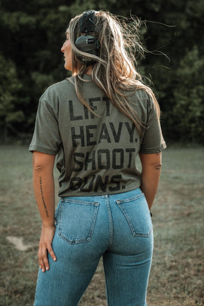 A woman with tattooed arms stands outdoors in jeans, wearing a green "Lift Heavy. Shoot Guns." shirt by Tango Charlie Apparel, made of polyester and combed ringspun cotton, with ear protection on her head, facing away toward trees and grass.