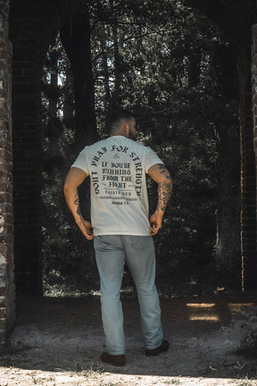 A man with tattooed arms stands under a brick archway, facing away, wearing the 13th Be Strong Premium Tee in white with printed text and light blue jeans. A forested area bathed in sunlight is visible in the background, lending an inspiring touch evocative of Joshua 1:9.