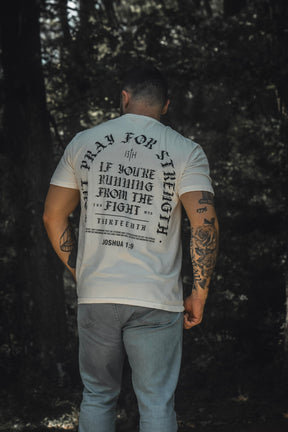 A man stands outdoors with his back to the camera, wearing the 13th Be Strong Premium Tee—a white shirt featuring a bold black message inspired by Joshua 1:9—and blue jeans. Trees and foliage are visible in the background.