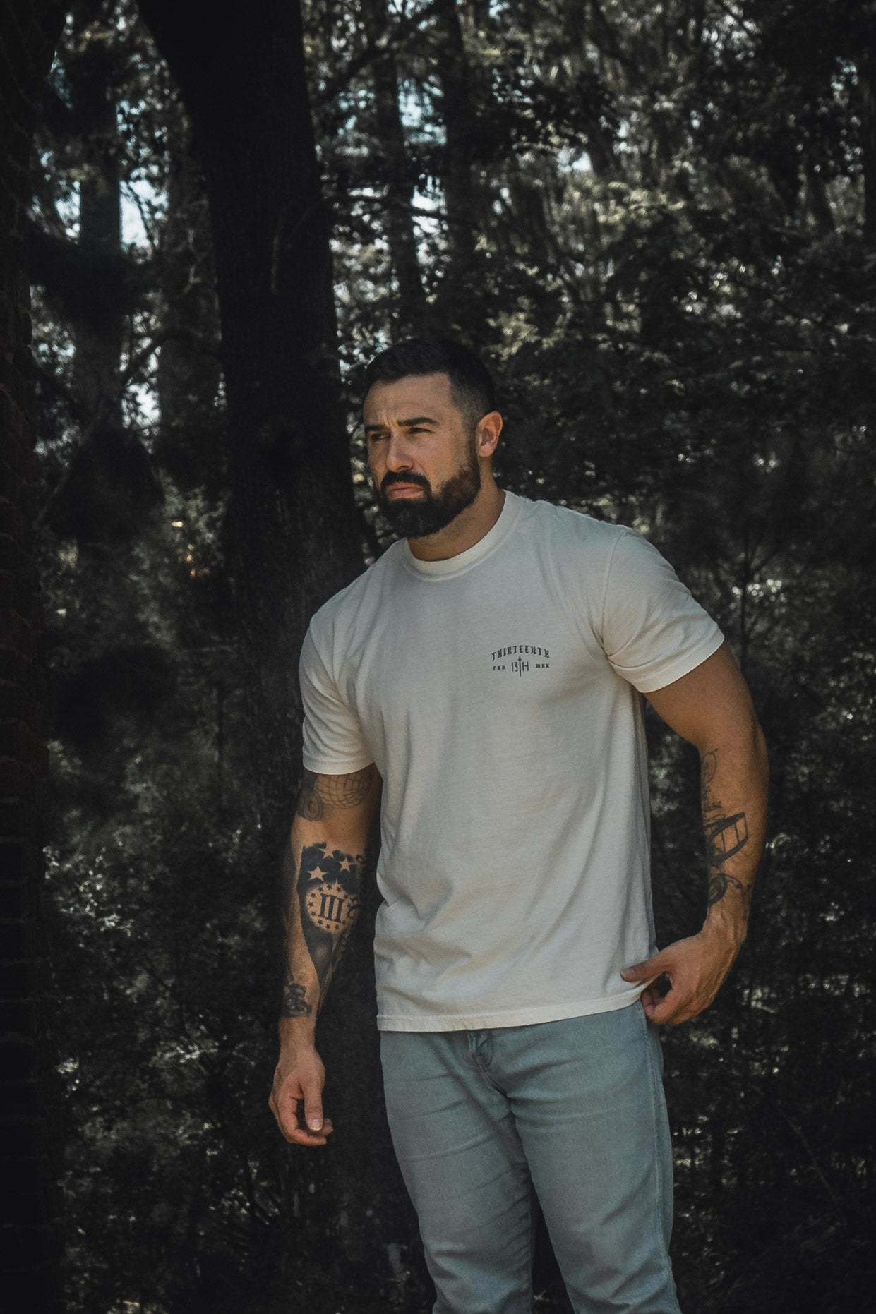 A tattooed man wearing the 13th Be Strong Premium Tee and light blue jeans stands outdoors in a wooded area, gazing to the side.