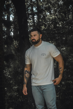 A tattooed man wearing the 13th Be Strong Premium Tee and light blue jeans stands outdoors in a wooded area, gazing to the side.