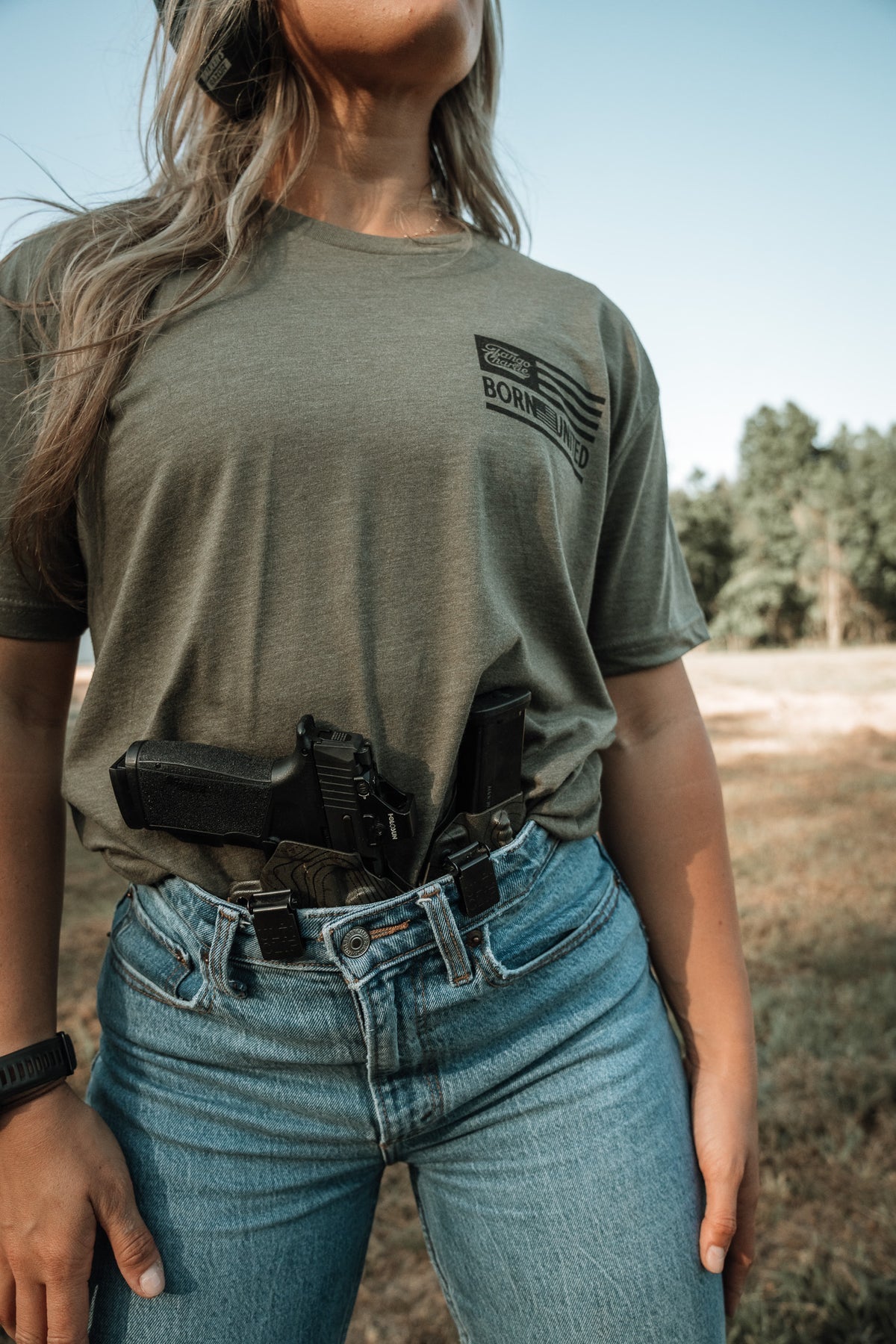 A person wearing a green "Lift Heavy. Shoot Guns." Next Level CVC shirt by Tango Charlie Apparel, tucked into blue jeans, stands outdoors with a holstered handgun. The soft cotton-poly blend ensures comfort amid the grassy, tree-lined scene.