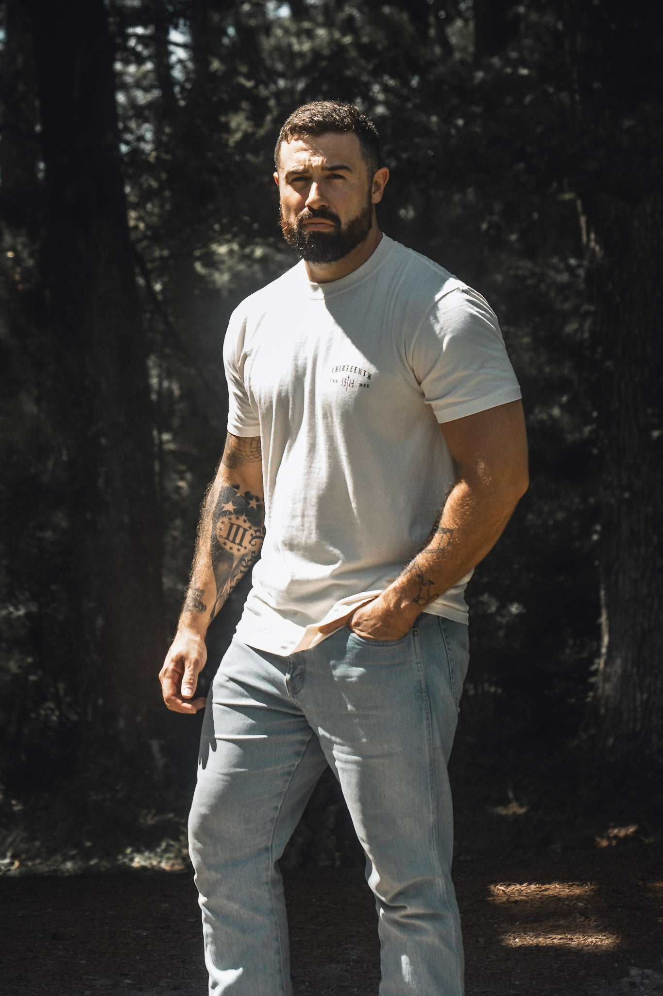 A bearded man stands outdoors in the sunlight, wearing the 13th Be Strong Premium Tee and jeans, with one hand in his pocket and trees in the background.
