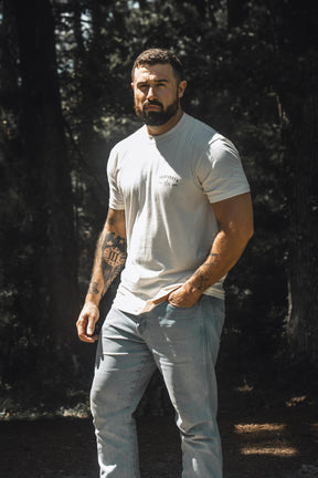A bearded man stands outdoors in the sunlight, wearing the 13th Be Strong Premium Tee and jeans, with one hand in his pocket and trees in the background.