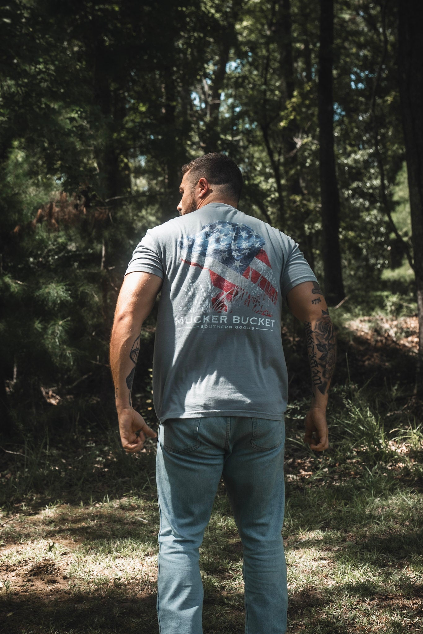 A man stands outdoors in a sunlit wooded area, facing away and wearing the Mucker Bucket Lab Flag Premium Tee—a relaxed fit shirt made from soft ring-spun cotton—featuring an American flag graphic and the words “MUCKER BUCKET” on the back.