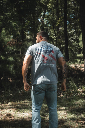 A man stands outdoors in a sunlit wooded area, facing away and wearing the Mucker Bucket Lab Flag Premium Tee—a relaxed fit shirt made from soft ring-spun cotton—featuring an American flag graphic and the words “MUCKER BUCKET” on the back.