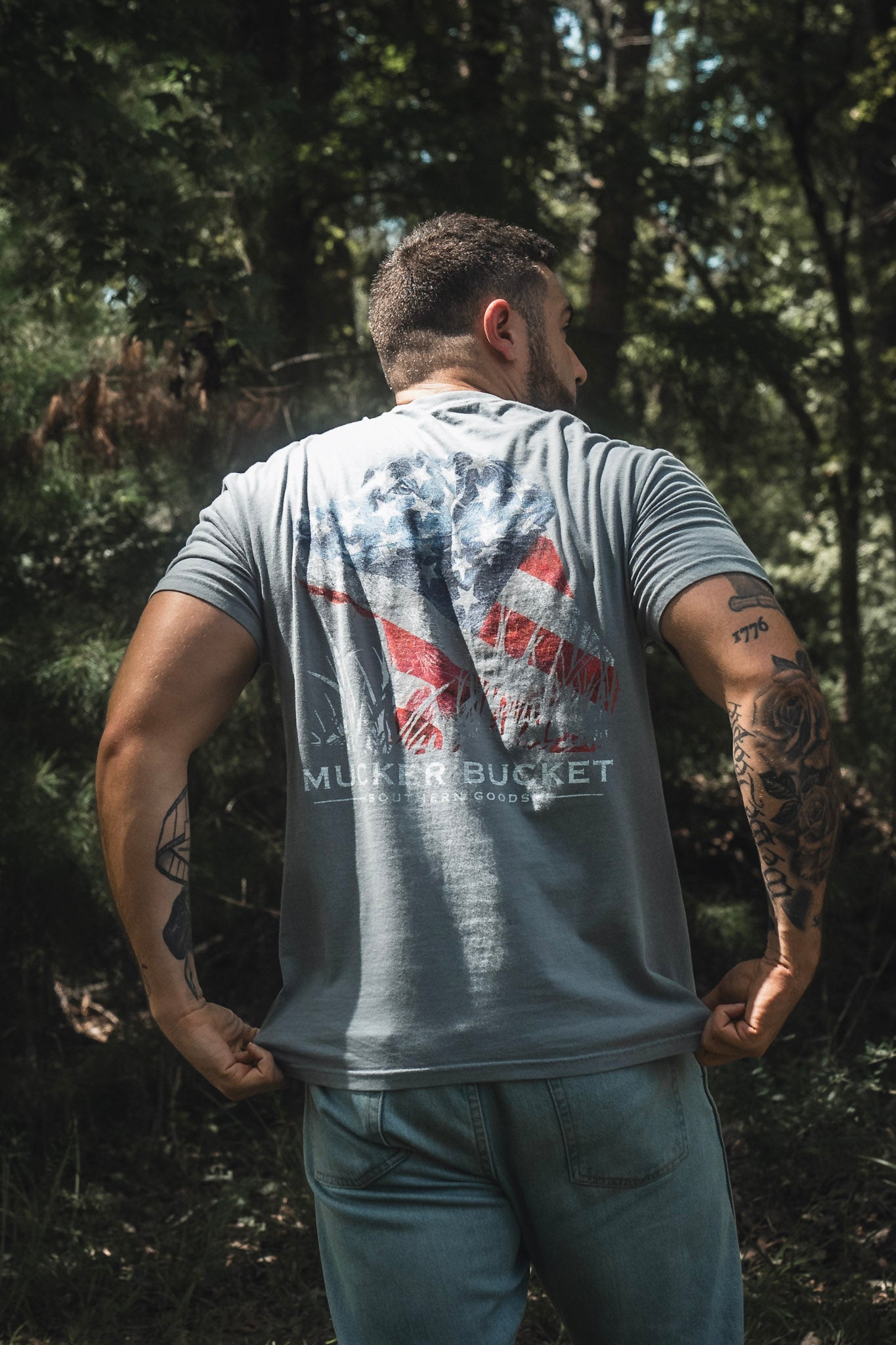 A man stands outdoors with his back to the camera, wearing the Mucker Bucket Lab Flag Premium Tee—a relaxed fit, gray shirt made of soft ring-spun cotton and featuring a faded American flag graphic. Paired with blue jeans, this premium tee is perfectly suited to the setting of sunlit trees in the background.