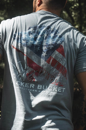 Outdoors, a person wears the Lab Flag Premium Tee by Mucker Bucket—a gray, premium relaxed fit shirt crafted from soft ring-spun cotton and featuring a large weathered Confederate flag graphic with partially visible BECKER BUCKET SOUTHERN GOODS text. This tee offers comfort and classic Southern style.