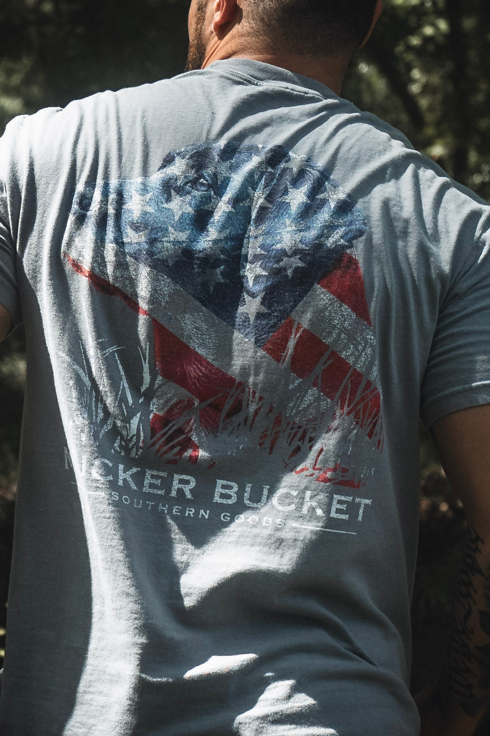 Outdoors, a person wears the Lab Flag Premium Tee by Mucker Bucket—a gray, premium relaxed fit shirt crafted from soft ring-spun cotton and featuring a large weathered Confederate flag graphic with partially visible BECKER BUCKET SOUTHERN GOODS text. This tee offers comfort and classic Southern style.