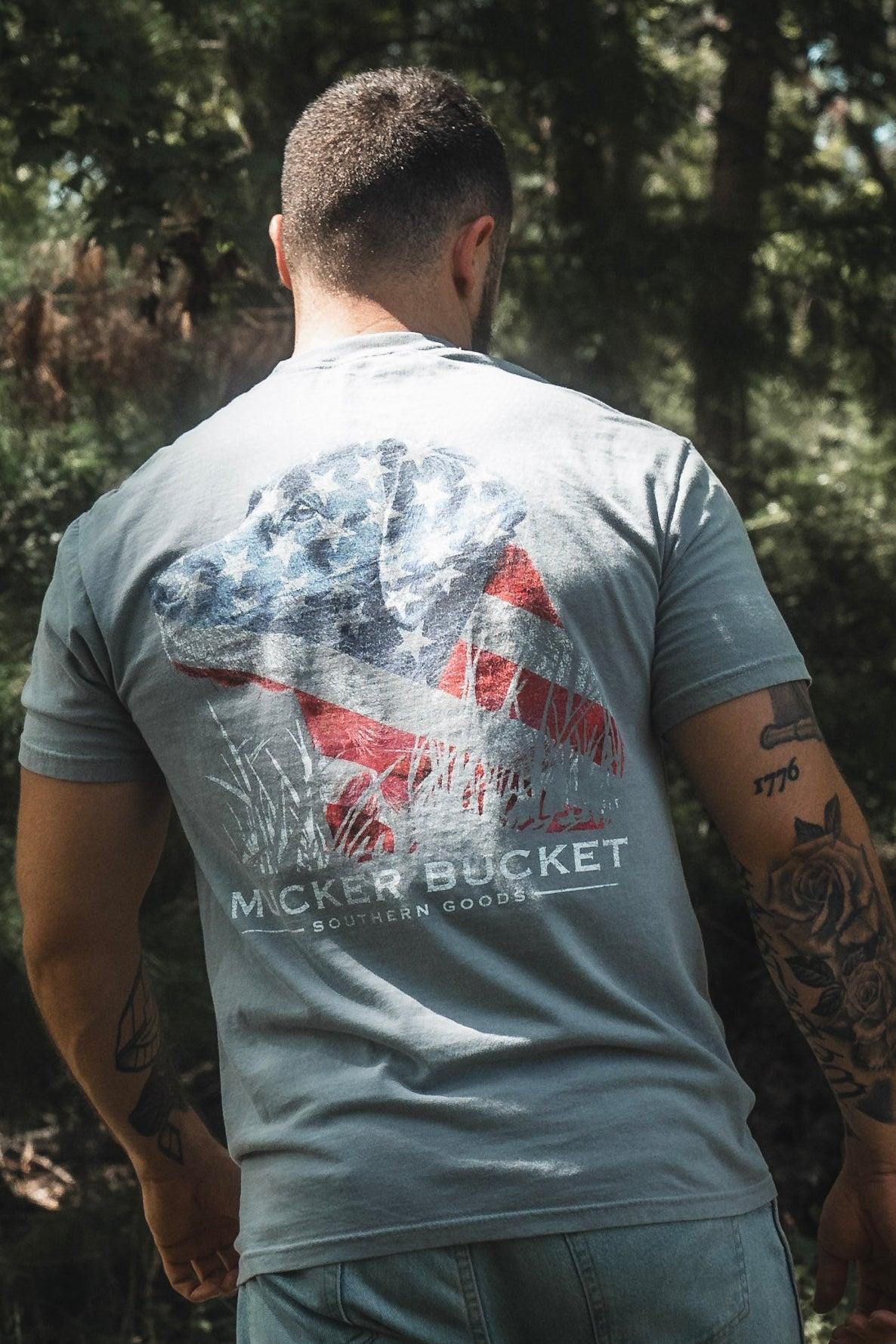 A man stands outdoors wearing the Mucker Bucket Lab Flag Premium Tee, a gray relaxed fit shirt made from soft ring-spun cotton. The large back graphic features a dog overlaid with an American flag design and the text “Mucker Bucket Southern Goods.”.
