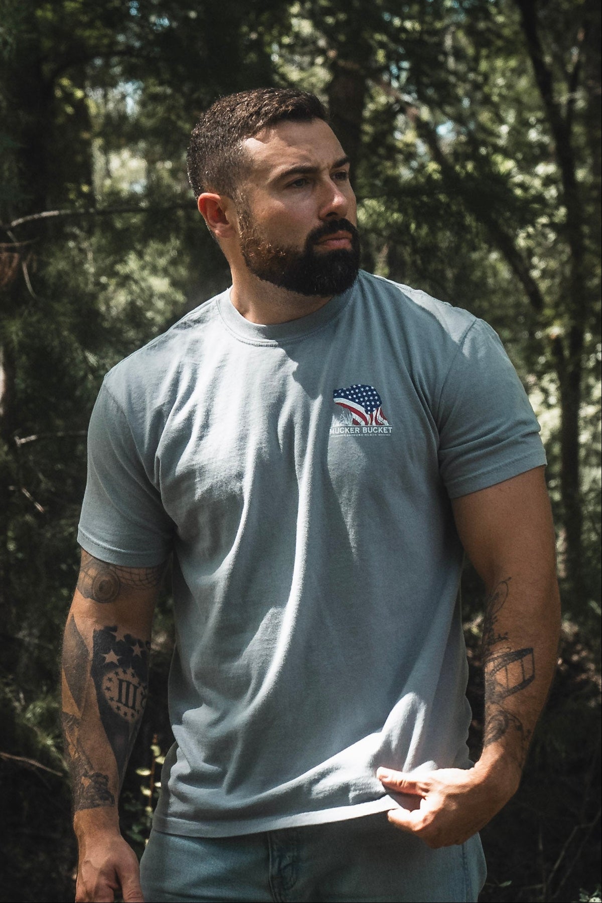 A bearded, tattooed man stands outdoors amidst trees and greenery, wearing the Mucker Bucket Lab Flag Premium Tee—a gray, relaxed fit shirt crafted from soft ring-spun cotton and featuring an American flag graphic. The natural setting emphasizes the comfort and timeless style of this premium tee.