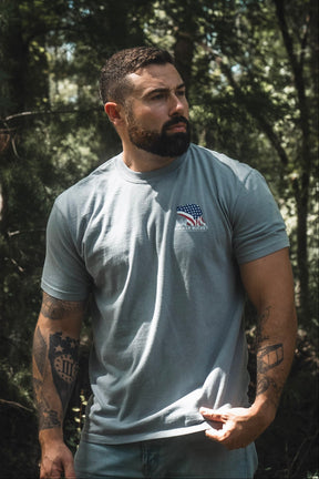 A bearded, tattooed man stands outdoors amidst trees and greenery, wearing the Mucker Bucket Lab Flag Premium Tee—a gray, relaxed fit shirt crafted from soft ring-spun cotton and featuring an American flag graphic. The natural setting emphasizes the comfort and timeless style of this premium tee.