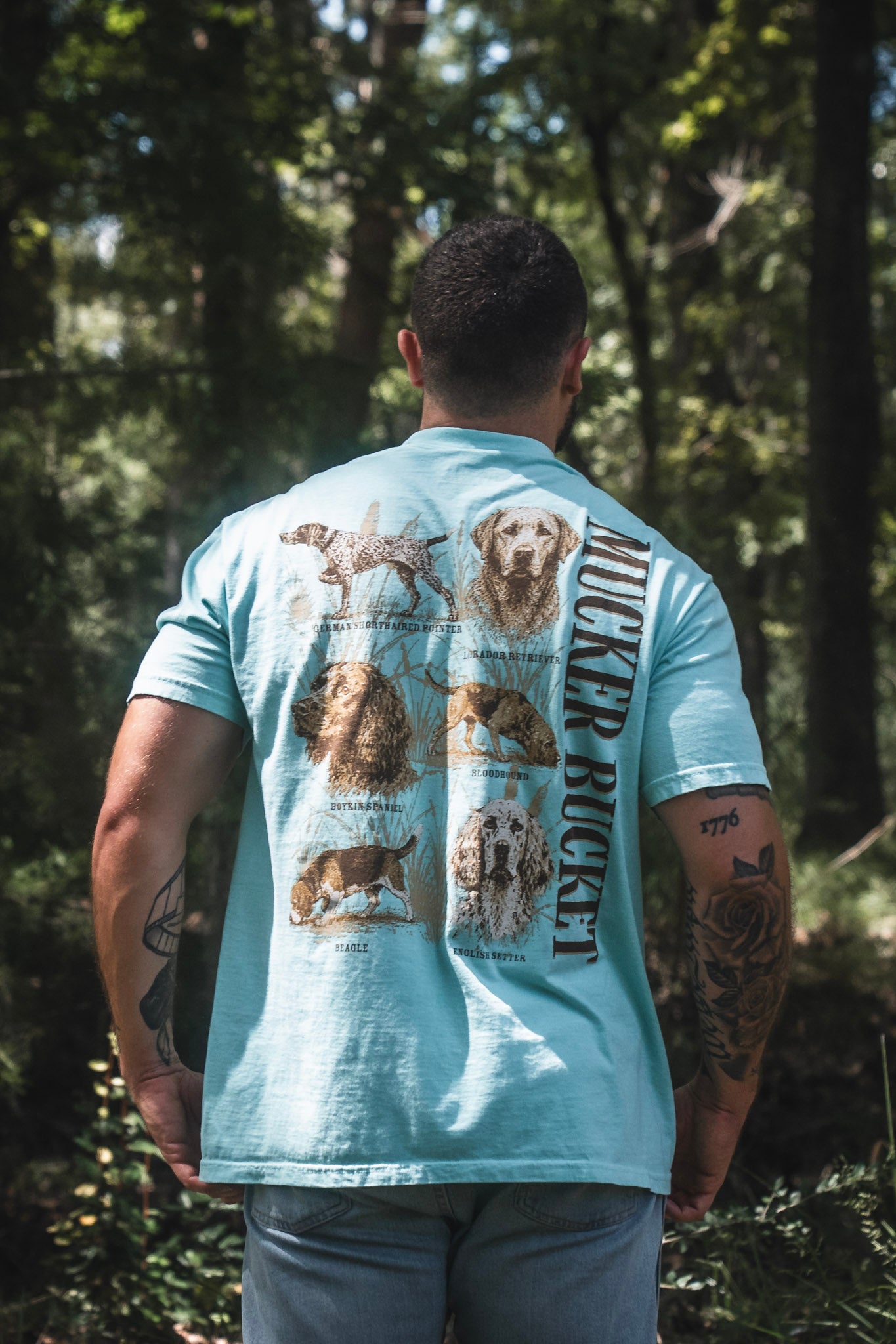 A person stands outdoors with their back to the camera, wearing the Mucker Bucket Dog DNA Premium Tee—a relaxed fit light blue shirt adorned with whimsical illustrations and names of different dog breeds on the back. This premium tee pops against a lush backdrop of trees and greenery, making it an ideal choice for nature lovers and pet enthusiasts alike.