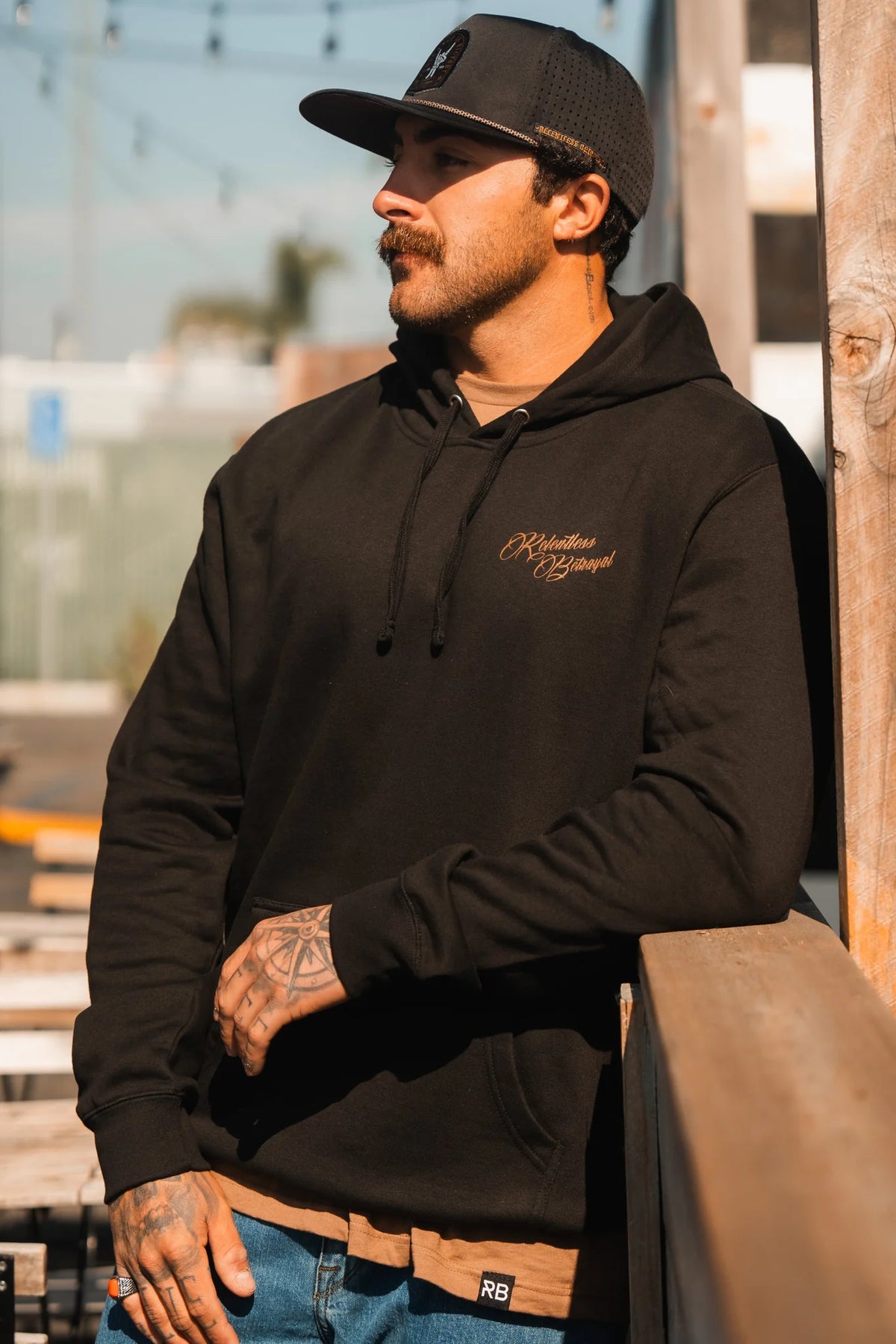 A tattooed man with a mustache leans on an outdoor wooden railing, wearing blue jeans, a black cap, and the Vengeance Hoodie by Relentless Betrayal. Sunlight illuminates his face as he gazes into the distance with resilience.