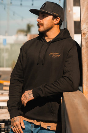 A tattooed man with a mustache leans on an outdoor wooden railing, wearing blue jeans, a black cap, and the Vengeance Hoodie by Relentless Betrayal. Sunlight illuminates his face as he gazes into the distance with resilience.