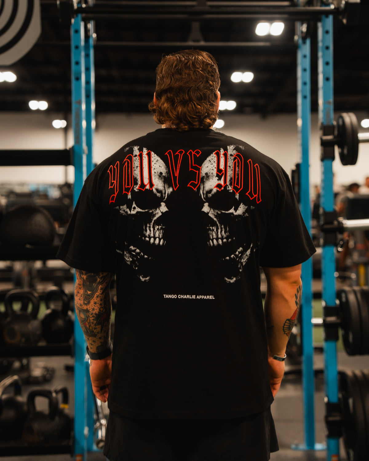 A man with tattooed arms stands in a gym wearing the Tango Charlie Apparel You vs You Premium Oversized Tee, featuring two skulls and bold red YOU VS YOU text. Gym equipment and weights are visible in the background.