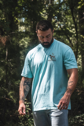 A man with tattoos stands outdoors in a wooded area, looking down with a relaxed posture while wearing the Mucker Bucket Dog DNA Premium Tee in light blue.