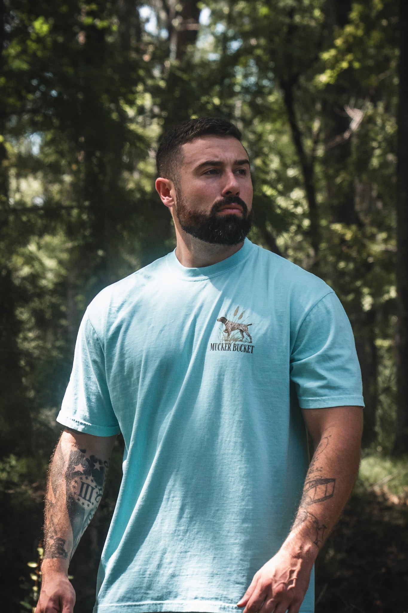 A bearded man with tattoos stands outdoors in a sunlit wooded area, wearing the Mucker Bucket Dog DNA Premium Tee in light blue with a relaxed fit.