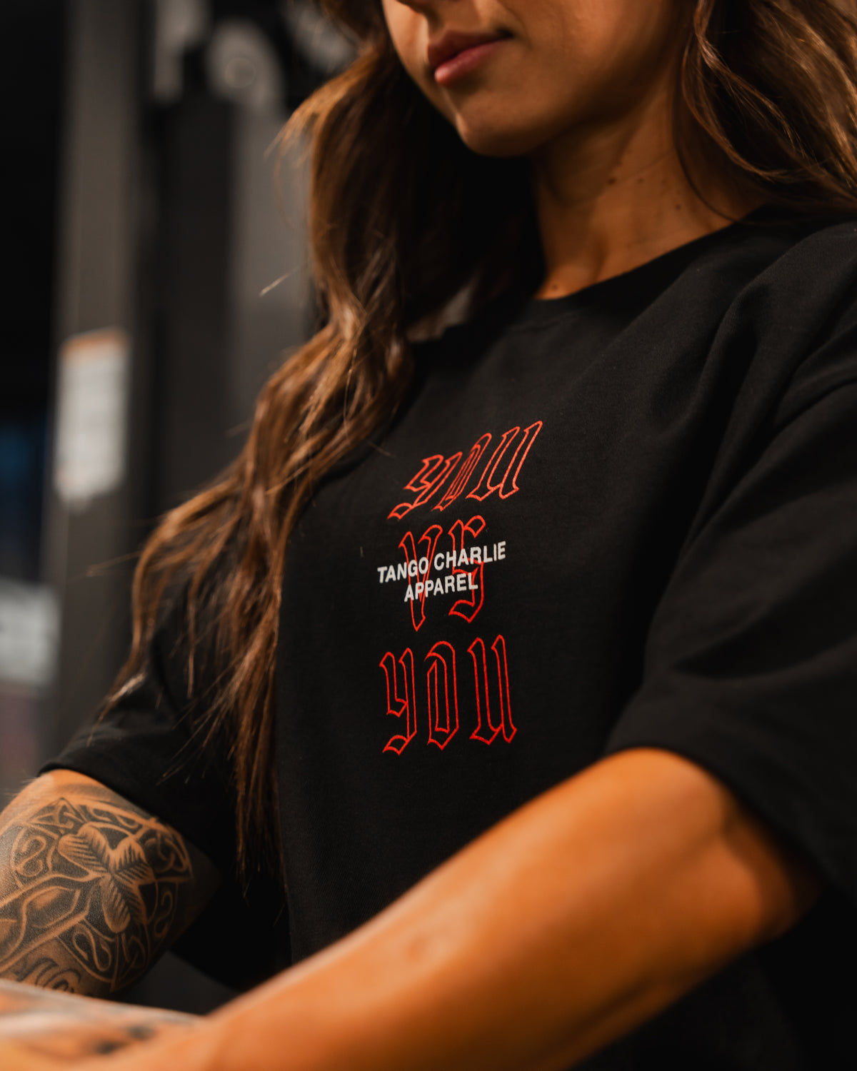 A woman with long brown hair and a tattooed arm wears the You vs You - Premium Oversized Tee by Tango Charlie Apparel. The focus is on her upper body and shirt, highlighting the tees premium heavyweight cotton quality.