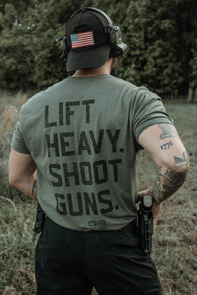Wearing the olive green Tango Charlie Apparel "Lift Heavy. Shoot Guns." tee, made from combed ringspun cotton and polyester, a person stands outside among trees and grass, styled with tattoos, a headset, flag patch cap, and pistol holster.