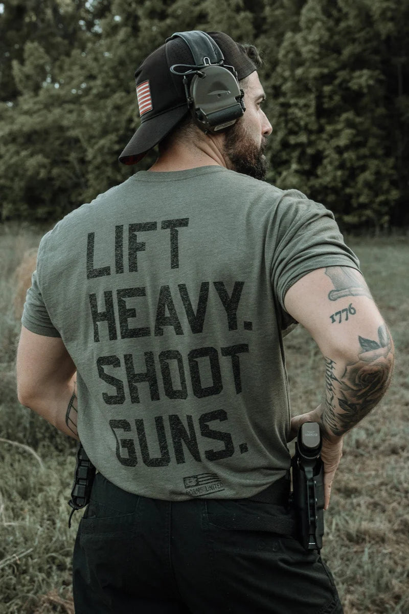A man outdoors, facing away with tattoos, holstered firearms, hat backward, and ear protection, wears the Tango Charlie Apparel "Lift Heavy. Shoot Guns." shirt. A forest is visible behind him.