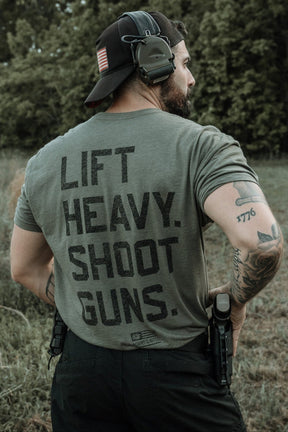 A tattooed man in a headset and cap stands outdoors, showing the back of his Tango Charlie Apparel "Lift Heavy. Shoot Guns." Next Level CVC Shirt—made from combed ringspun cotton and polyester—with a holstered gun amid greenery.