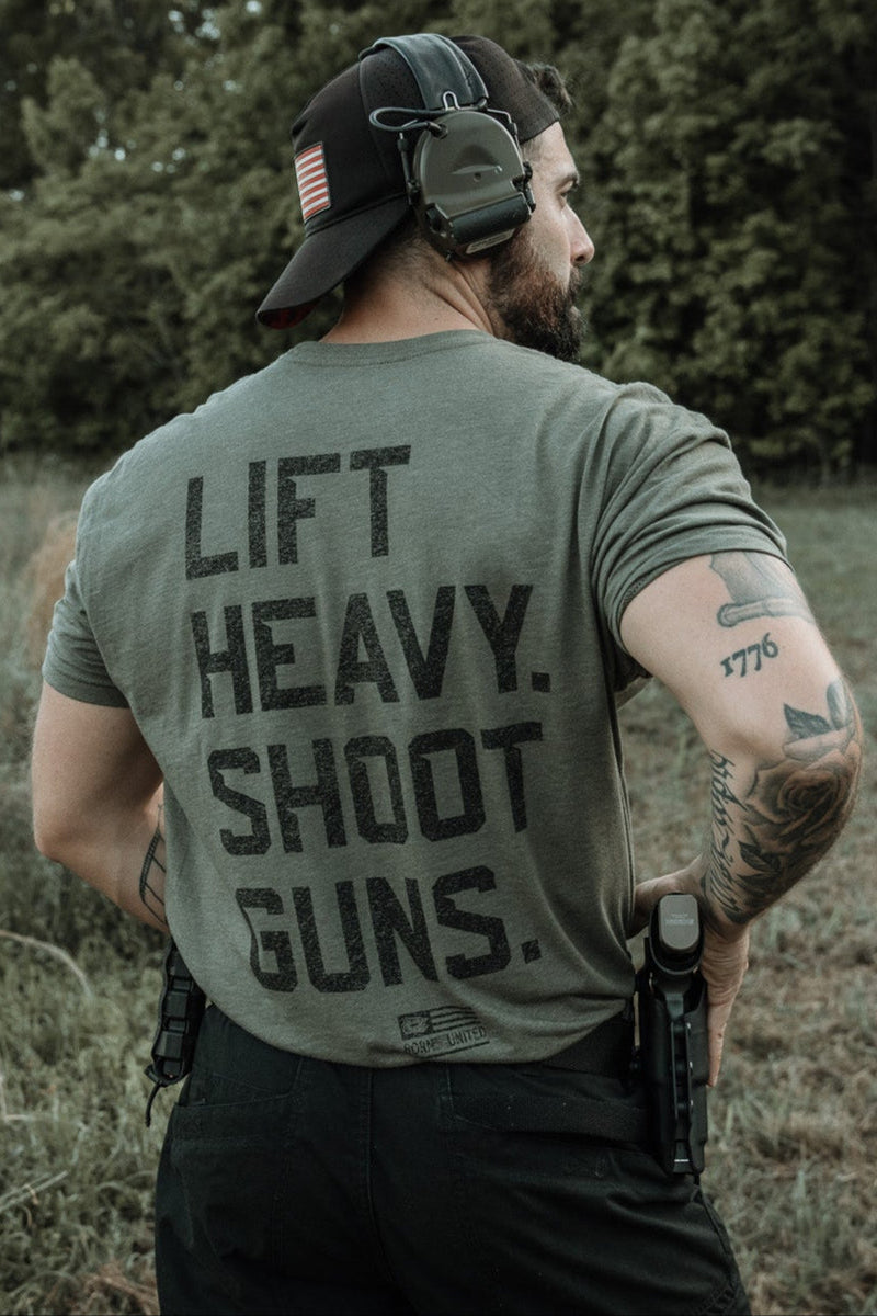 A tattooed man in a headset and cap stands outdoors, showing the back of his Tango Charlie Apparel "Lift Heavy. Shoot Guns." Next Level CVC Shirt—made from combed ringspun cotton and polyester—with a holstered gun amid greenery.